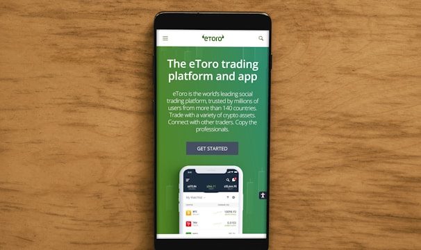 A smartphone displaying an advertisement for an online trading platform called eToro. The screen shows a green background with text promoting the platform as a leading social trading platform. A button labeled 'Get Started' is visible, along with a partial view of a mobile app screen below.